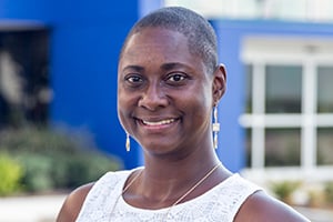 Smiling Black woman with short hair wearing a white sleeveless top, outdoors near a blue building.