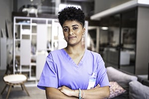 Healthcare professional wearing lavender scrubs stands with arms crossed in a modern clinic.