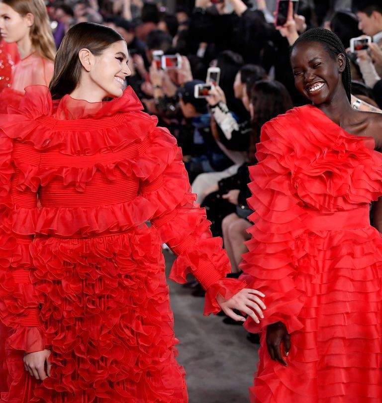 Two models in matching red ruffled gowns smiling at a fashion show, photographers in the background.