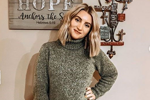 Woman with blonde hair wearing a gray knit turtleneck sweater, standing indoors with wall art and a hanging cross behind her.