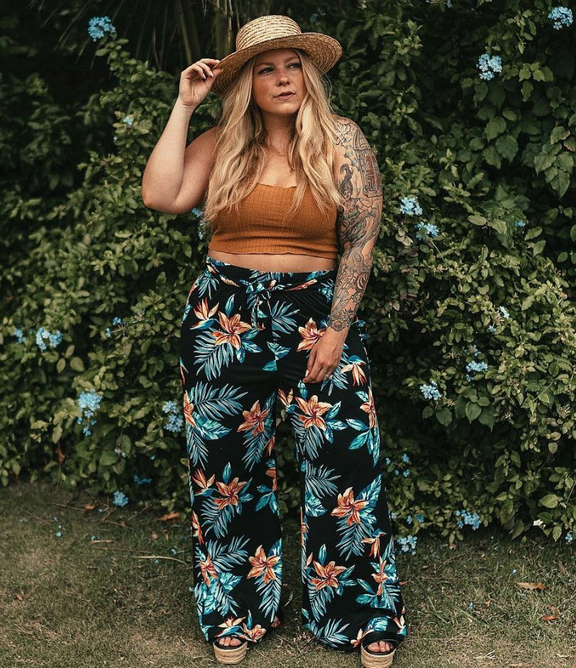 Woman in straw hat, rust-orange crop top, and tropical palm-leaf patterned wide-leg pants stands before a green hedge.
