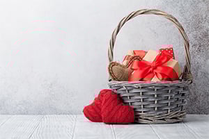 Wicker basket with red-wrapped gifts and a red heart-shaped yarn decoration beside it on a light wooden surface.