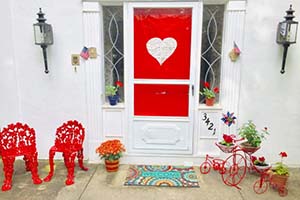 Front porch with red Valentine decor: red door panel with white heart, red metal chairs, potted plants, and pink plant cart.