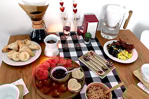 Brunch spread on a checked tablecloth with a coffee pot, bread, watermelon slices, cheese, nuts, and heart decorations.