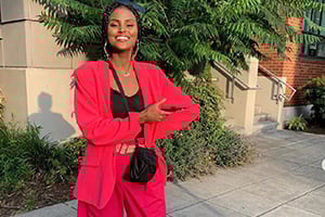 Smiling woman wearing a bright pink suit holds a red folder outdoors near a building.