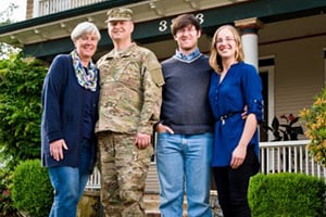 Family of four posing on a front porch, with a soldier in camouflage standing between two smiling adults.