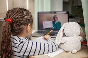 Child with braided hair studies at home, writing at a desk while a teacher appears on a laptop screen during a video call.