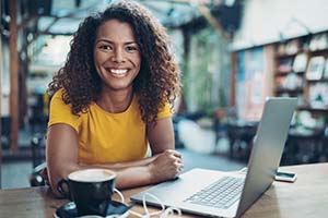 Smiling woman in a yellow shirt sits at a cafe table with a laptop.