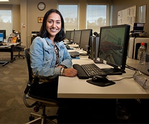 Smiling woman sits at a desk in an office with multiple computer monitors.