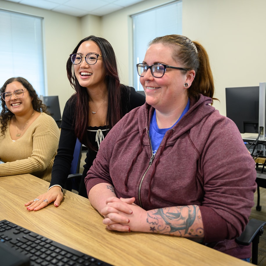 Three women smiling while collaborating at a desk with computers in a bright training room.