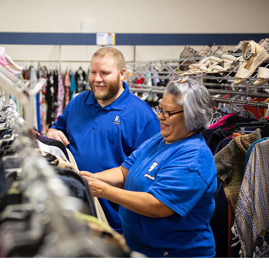 Two staff members in matching blue polo shirts assist a shopper among clothing racks in a thrift store.