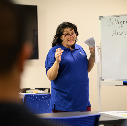 Instructor in a blue polo shirt teaching in a classroom, holding papers and gesturing toward students.