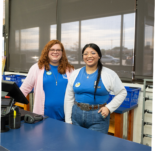 Two smiling women staff members stand behind a blue checkout counter in a store or classroom, wearing blue shirts and badges.