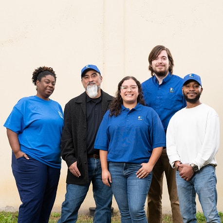 Five diverse coworkers standing outdoors against a light beige wall, wearing blue shirts with logos.