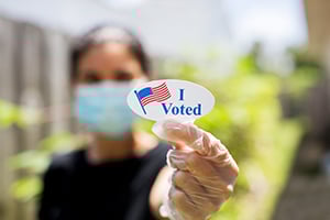 Close-up of a gloved hand holding an 'I Voted' sticker with a masked voter blurred in the background.