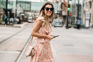 Woman in a peach floral dress, sunglasses, and crossbody bag walks on a city street while holding a smartphone.