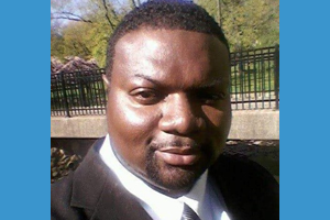 Close-up of a man in a dark suit and white tie outdoors, with trees and a fence in the background.