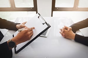 Two professionals sit across a white table in a meeting; one writes on a clipboard while the other clasps hands.