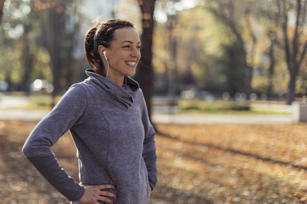 Smiling pregnant woman standing in a sunlit autumn park wearing a gray hoodie and earphones.