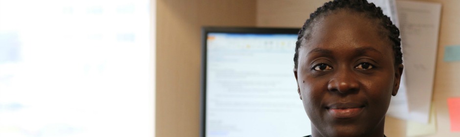 Professional woman with braided hair in an office, seated near a computer monitor.