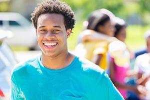 Smiling young person in a blue shirt outdoors with a blurred group in the background.