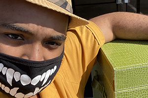 Close-up of a person wearing a hat and a black mask with white teeth print leaning on a green textured box.
