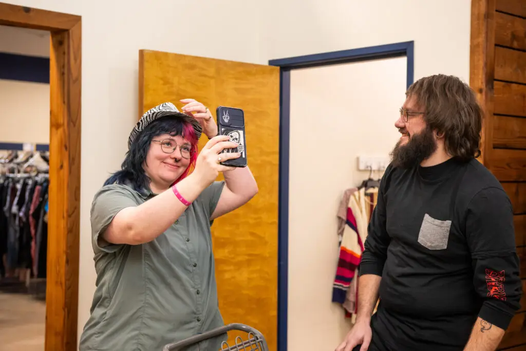Person with colorful hair and glasses takes a selfie with a phone in a clothing store while a bearded man smiles nearby.