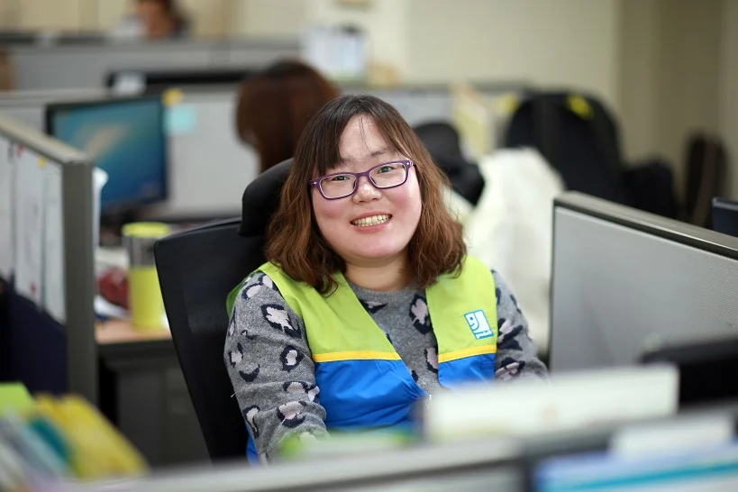 Smiling woman wearing a high-visibility vest sits at a desk in an open-plan office.