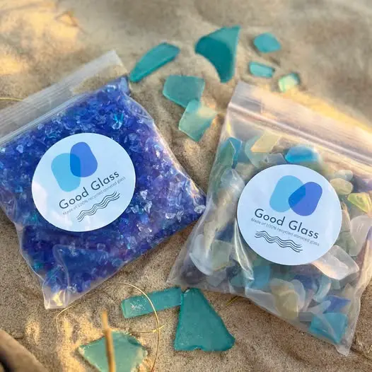 Two resealable bags of colorful sea glass labeled Good Glass on a sandy surface.