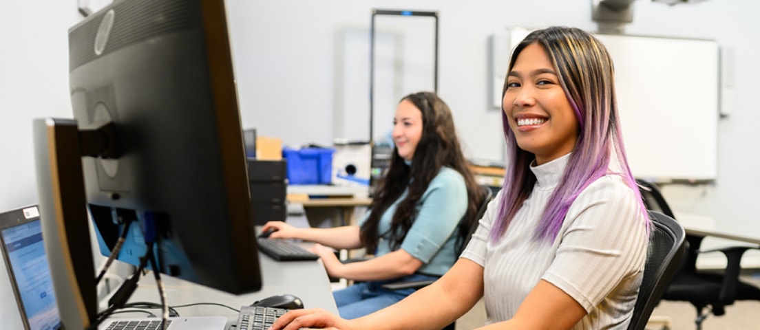Smiling woman with purple-tinted hair sits at a computer in an office, with a coworker working at a desk in the background.