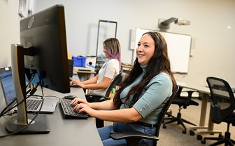 Smiling woman working at a computer in a modern classroom, with another student in the background