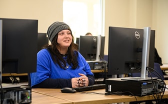 Student wearing a blue sweater and beanie sits at a computer desk in a lab, smiling.