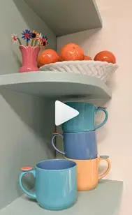 A white bowl holding oranges and three blue mugs and one yeallow on a shelf 
