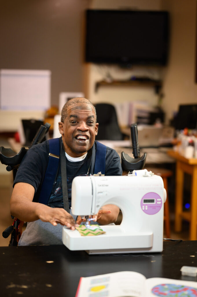 Smiling man in a wheelchair sewing fabric at a sewing machine in a workshop.