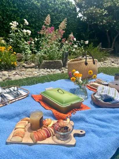 An image of a pinic setup in a garden with a purse, a cheese and crackers tray and flowers on top of a blue blanket